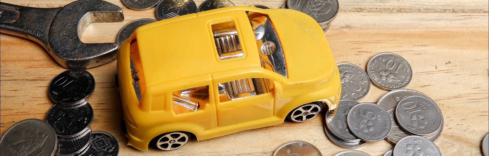 Gul leksaksbil i plast parkerad på furubord med verktyg och internationella mynt strödda omkring sig.